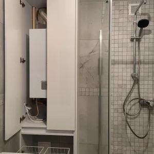 Biała zabudowa łazienkowa na wymiar z ukrytą instalacją i szafką techniczną obok kabiny prysznicowej.