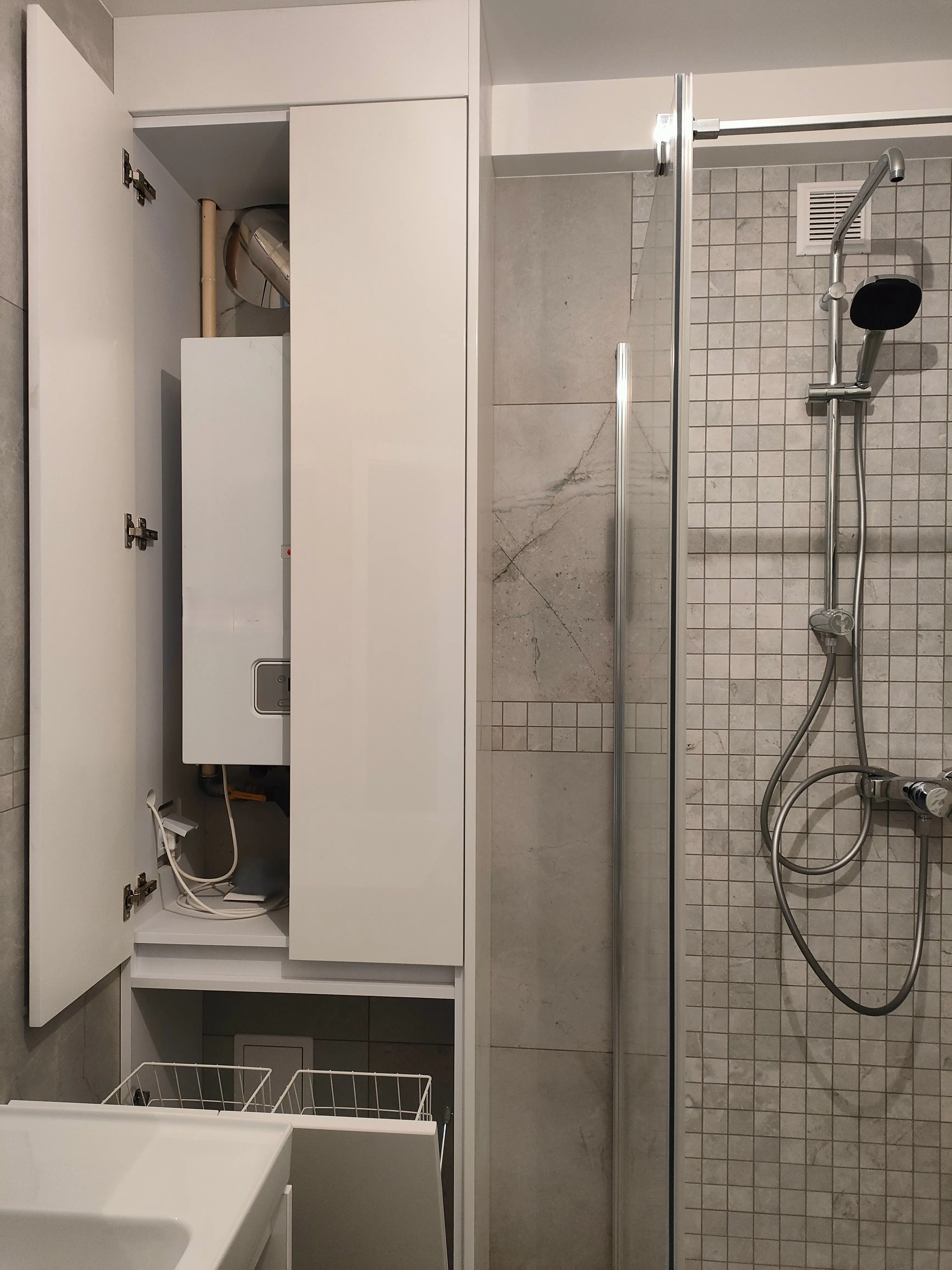 Biała zabudowa łazienkowa na wymiar z ukrytą instalacją i szafką techniczną obok kabiny prysznicowej.